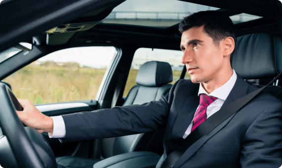 A chauffeur in a dark suit with a red tie driving through a scenic route, viewed from the passenger seat.