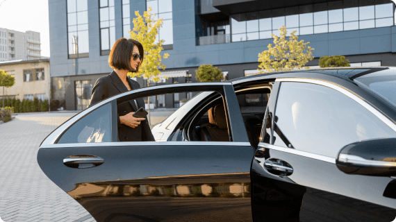 Confident woman in black suit about to step into a sleek black luxury car, captured from a sunny side angle near modern office buildings.