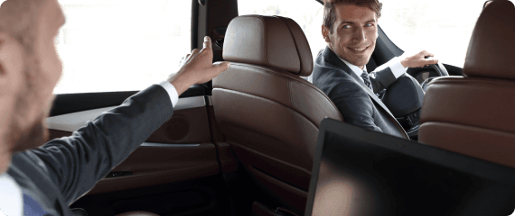 Smiling chauffeur in a suit engages with a suited passenger seated in the back of a luxury vehicle with tan interiors.