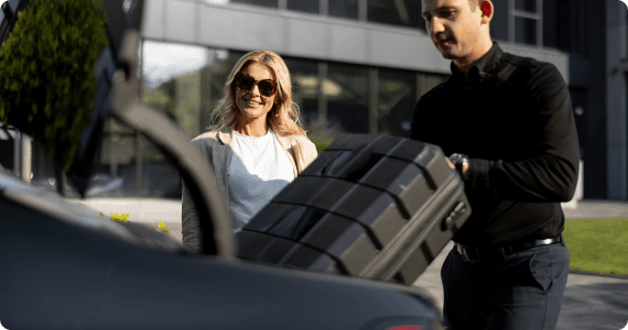 A chauffeur loads a black hard-shell suitcase into a trunk as a woman in sunglasses smiles nearby outside a modern building.