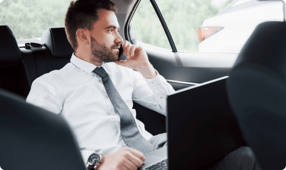 Businessman making a call while working on a laptop in the backseat of a luxury sedan.