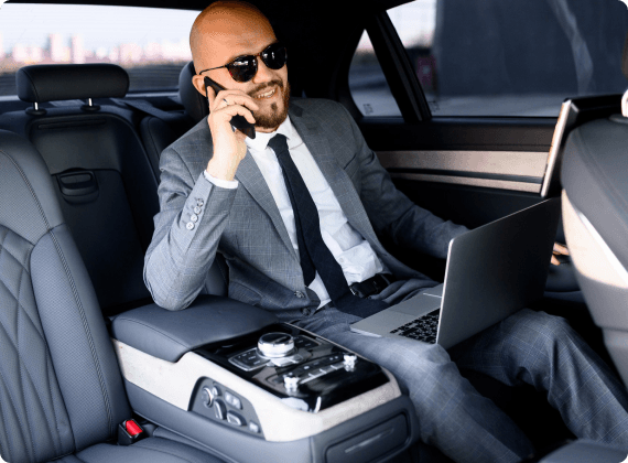 Business executive in a tailored grey suit, seated in a luxury car's rear seat with a laptop and phone, exuding productivity on the go.