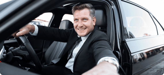 A smiling chauffeur leans from the driver’s seat of a black luxury car, inviting the guest aboard.