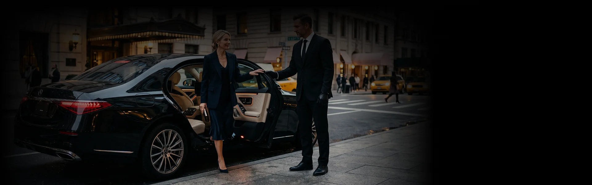 A professional chauffeur in a dark suit and gloves holds open the door of a sleek black luxury sedan parked on a city street. A businesswoman in a dark suit steps out of the vehicle onto the sidewalk in front of a classic city building.
