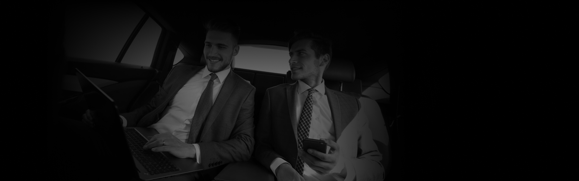 Two businessmen in suits work and chat inside a luxury backseat, showcasing comfort and flexibility of an Hourly Hire Service from a side-angle view.