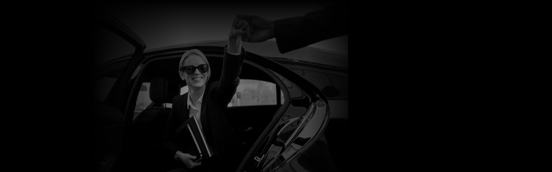 Smiling woman in sunglasses stepping out of a luxury car and reaching for a handshake, showcasing point-to-point car service with a door-side perspective.