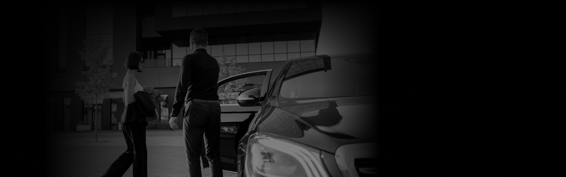 A man opens the door of a sleek black luxury car for a woman dressed elegantly, set against a modern building backdrop, ideal for Special Events.